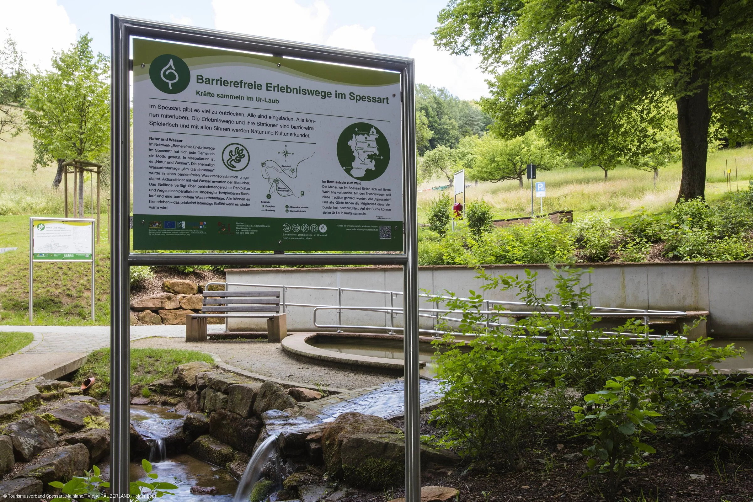 Ein Schild beschreibt eine barrierefreie Wassertretanlage, im Hintergrund sind das Becken, Wiesen und Bäume zu sehen