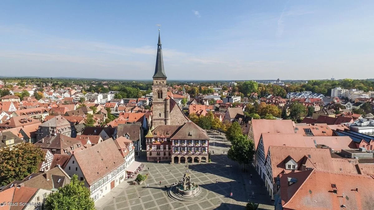 Eine Kirche vor einem Platz, in dessen Mitte ein Brunnen steht, ringsherum sind die Häuser einer Altstadt mit roten Dächern