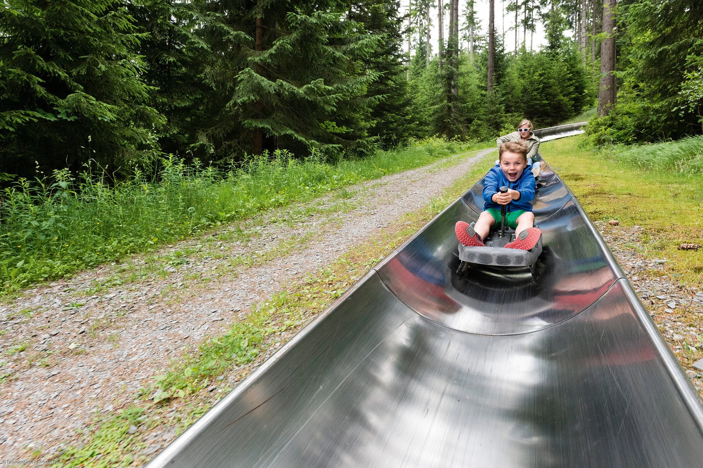 Zwei Kinder sausen hintereinander mit freudig erregten Gesichtern eine Sommerrodelbahn hinunter