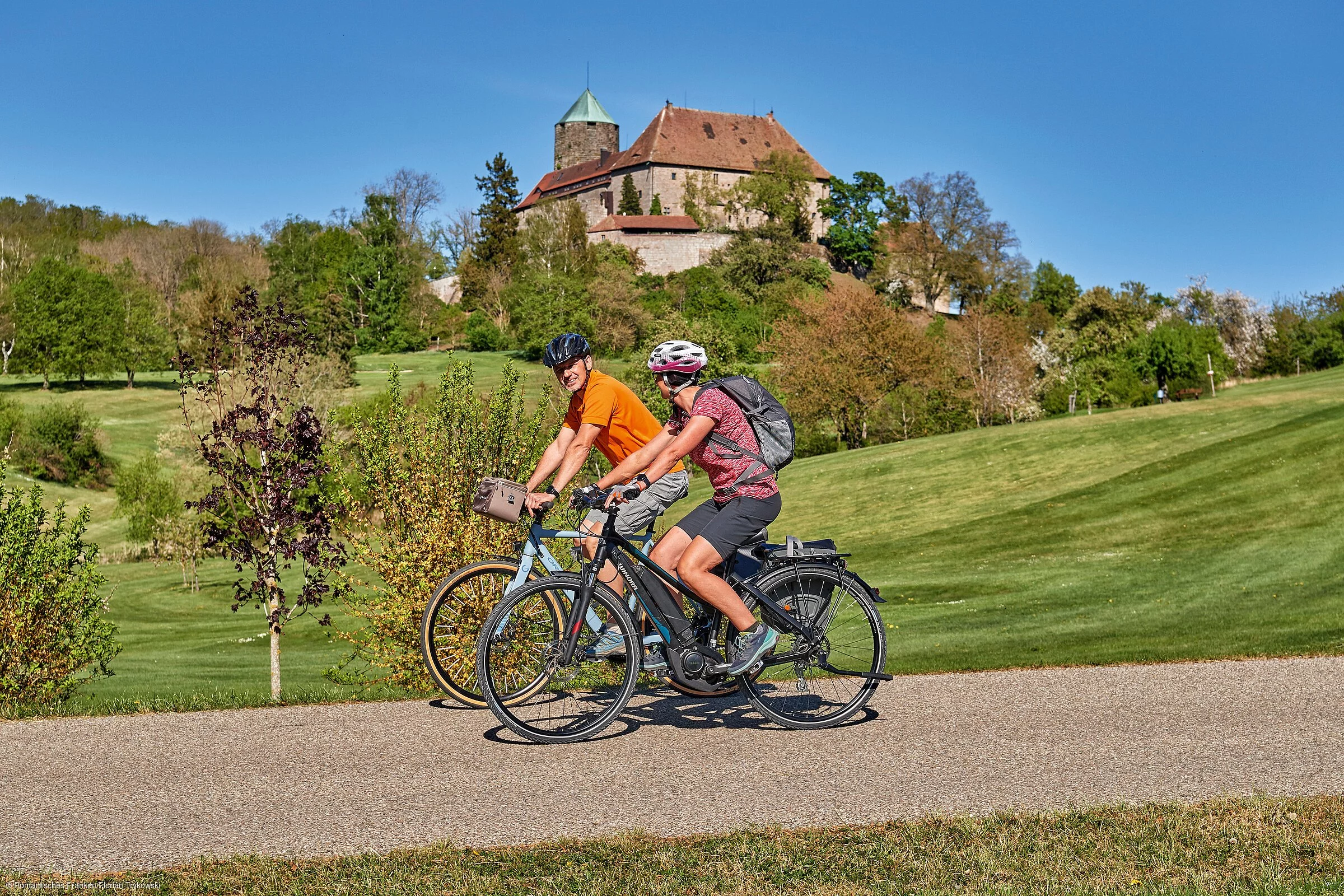 Zwei Radfahrer fahren bei schönem Wetter unterhalb einer Burg entlang