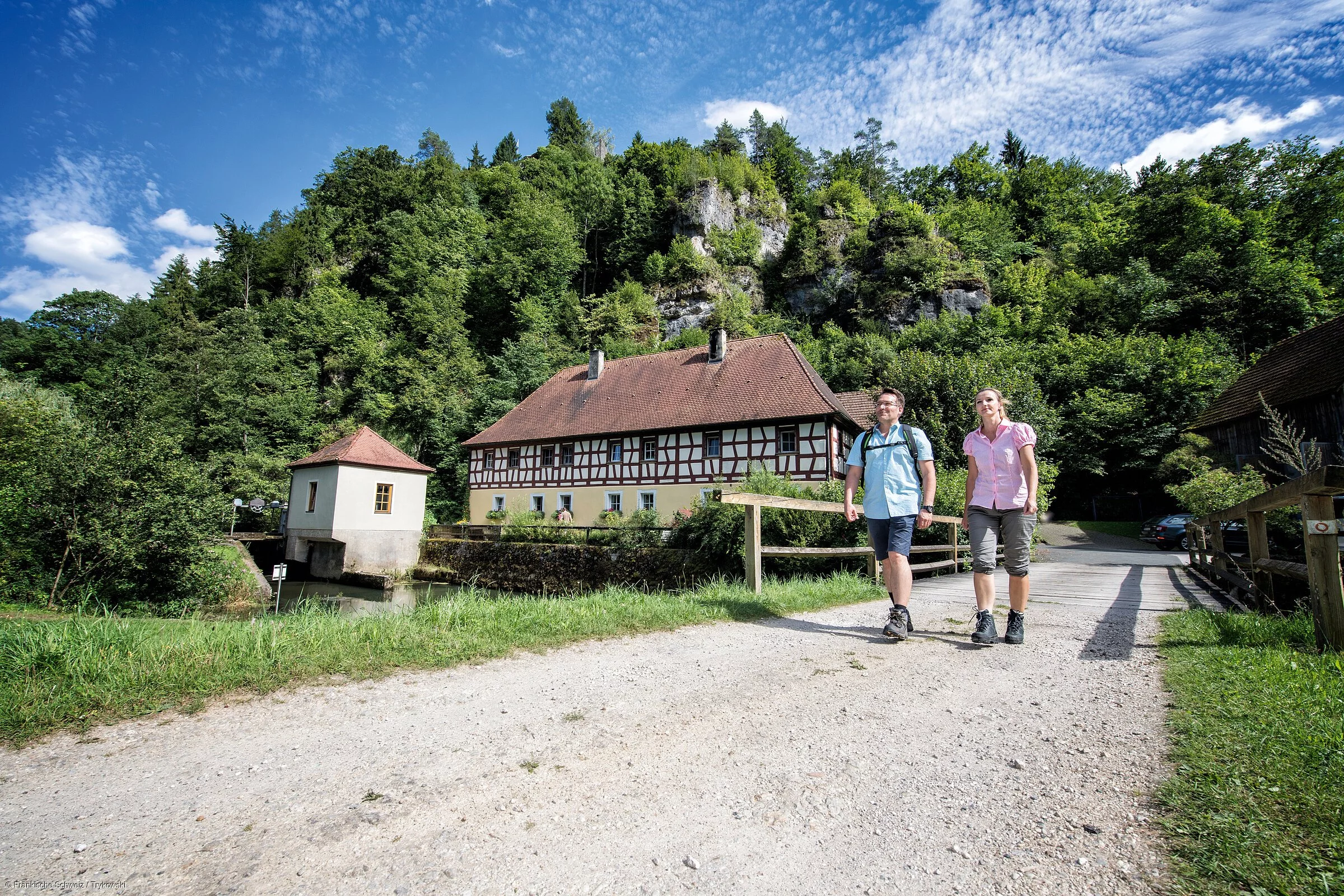 Auf einem breiten Schotterweg gehen zwei Wanderer vorbei an einem historischen Fachwerkgebäude