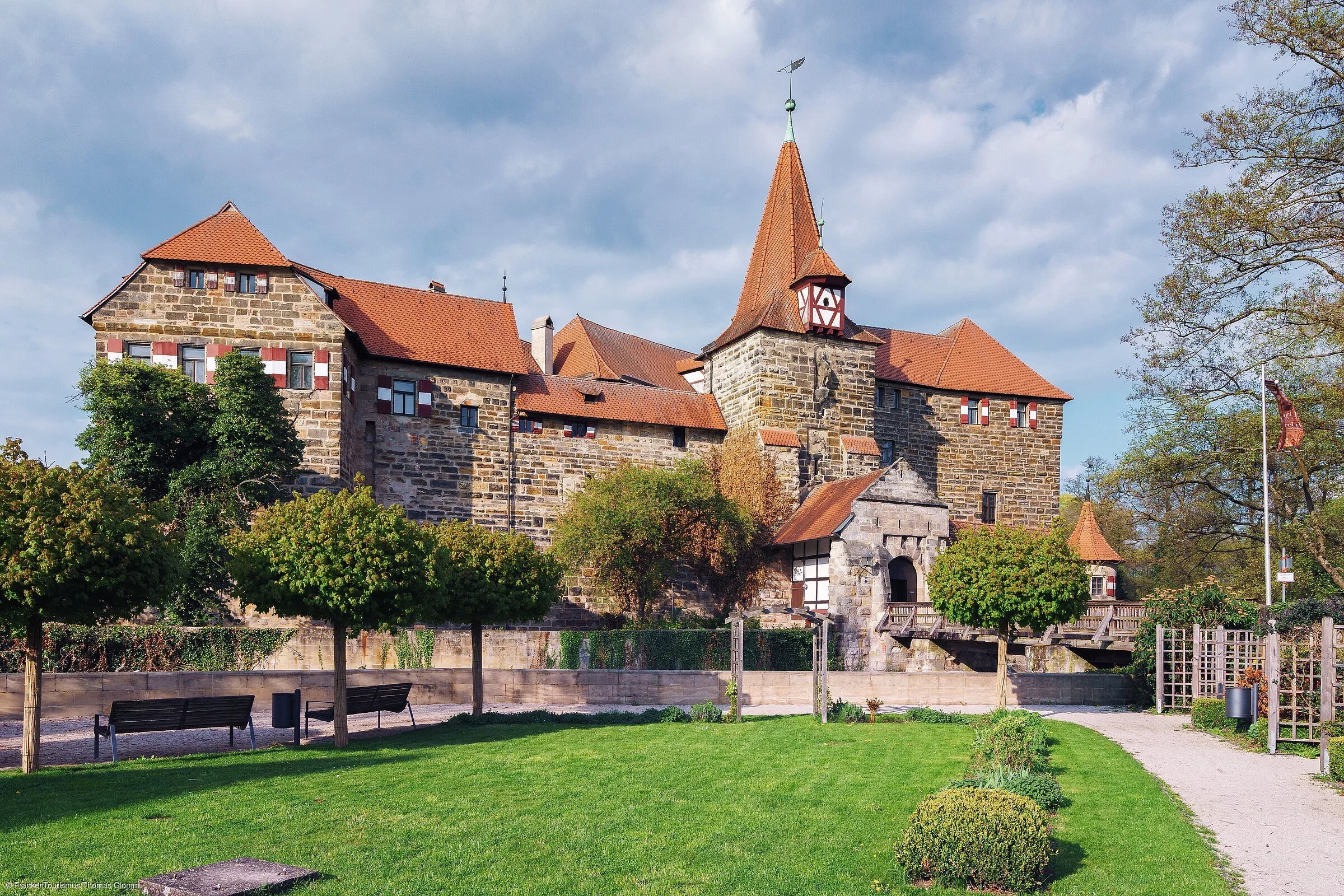 Eine mächtige Burg mit roten Dächern, davor eine grüne Wiese und Wege, die zur Burg führen