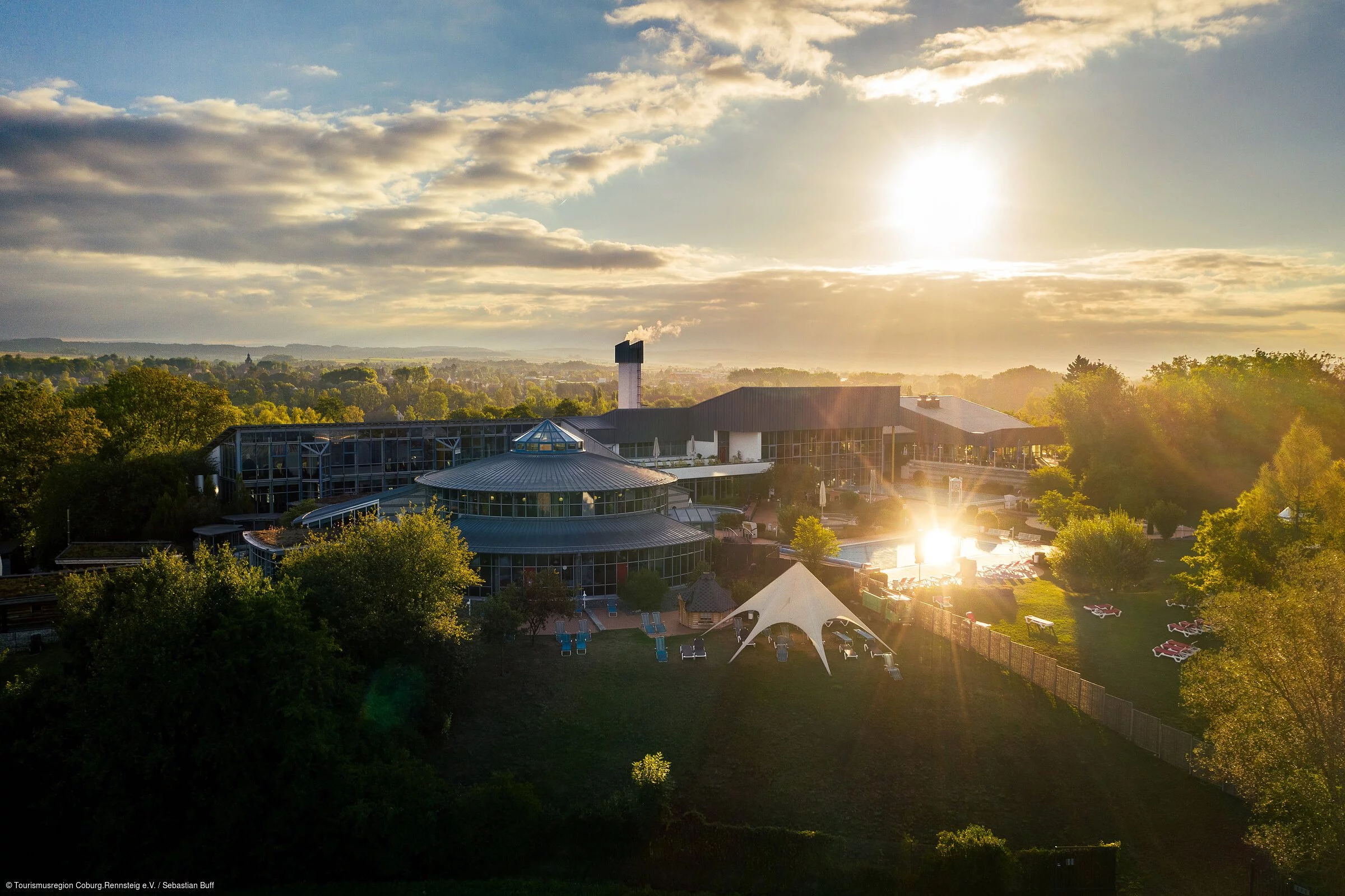 Eine Therme inmitten von Bäumen, im Hintergrund die morgendliche, aufgehende Sonne