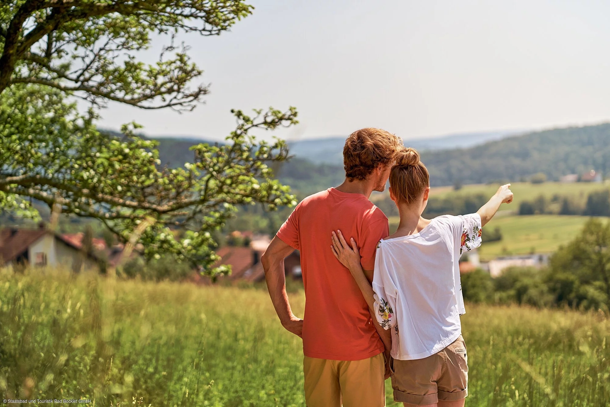 Ein Pärchen steht sommerlich gekleidet leicht erhöht und schaut in die Hügel und Felder vor ihnen