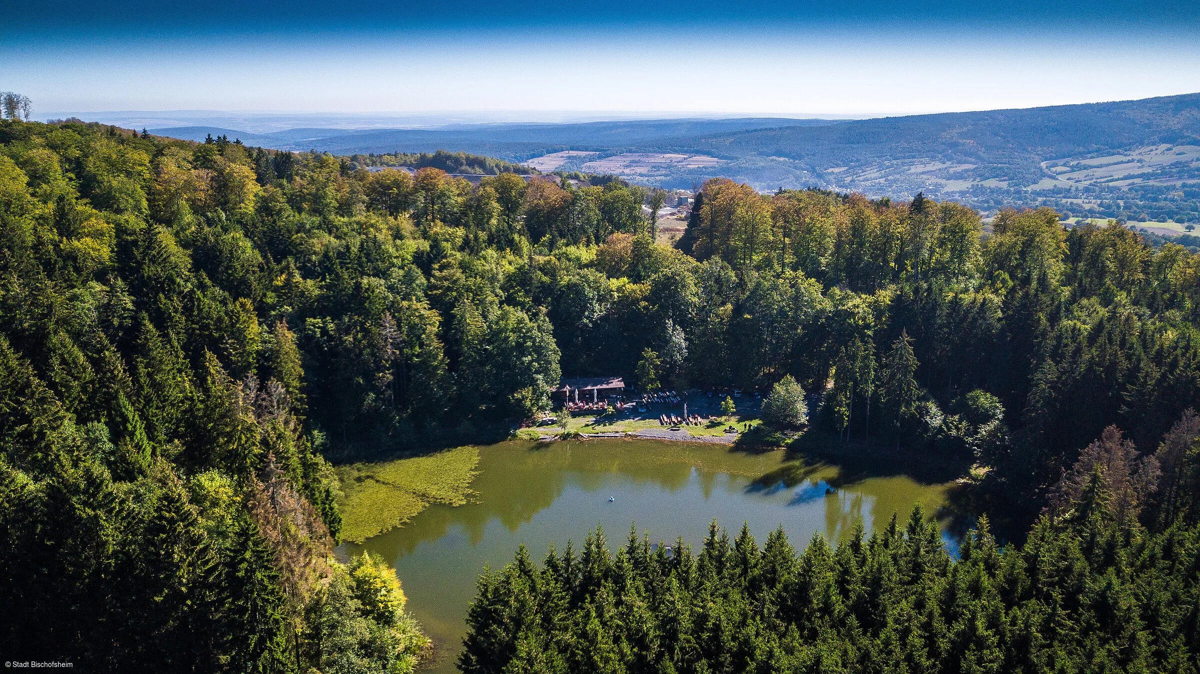 Ein Luftbild von einem kleinen See inmitten eines Nadelwaldes, im Hintergrund der Fernblick über bewaldete Hügel