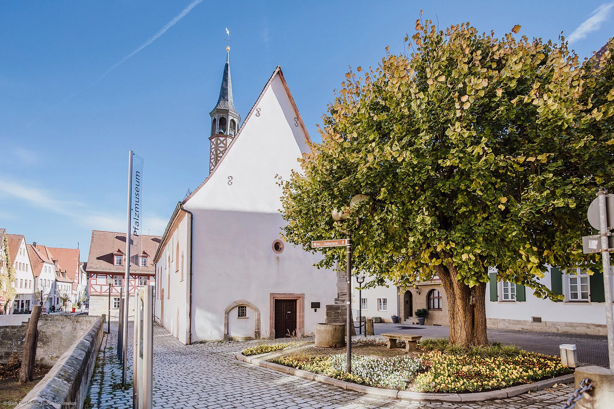 Eine kleine Kapelle in einer Stadt, daneben ein Bach und ein grün blühender Baum