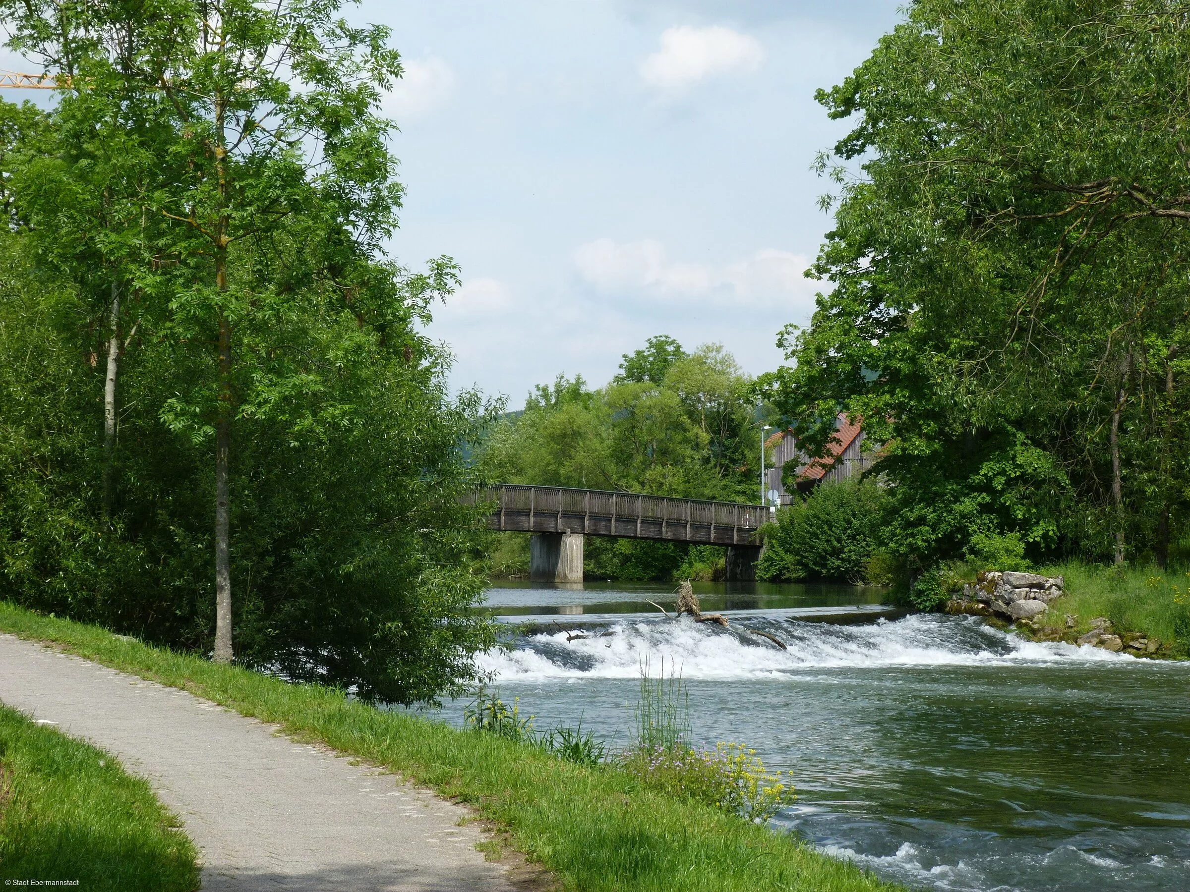 Neben einem kleinem Wehr in einem Fluss verläuft ein Weg, im Hintergrund eine Brücke