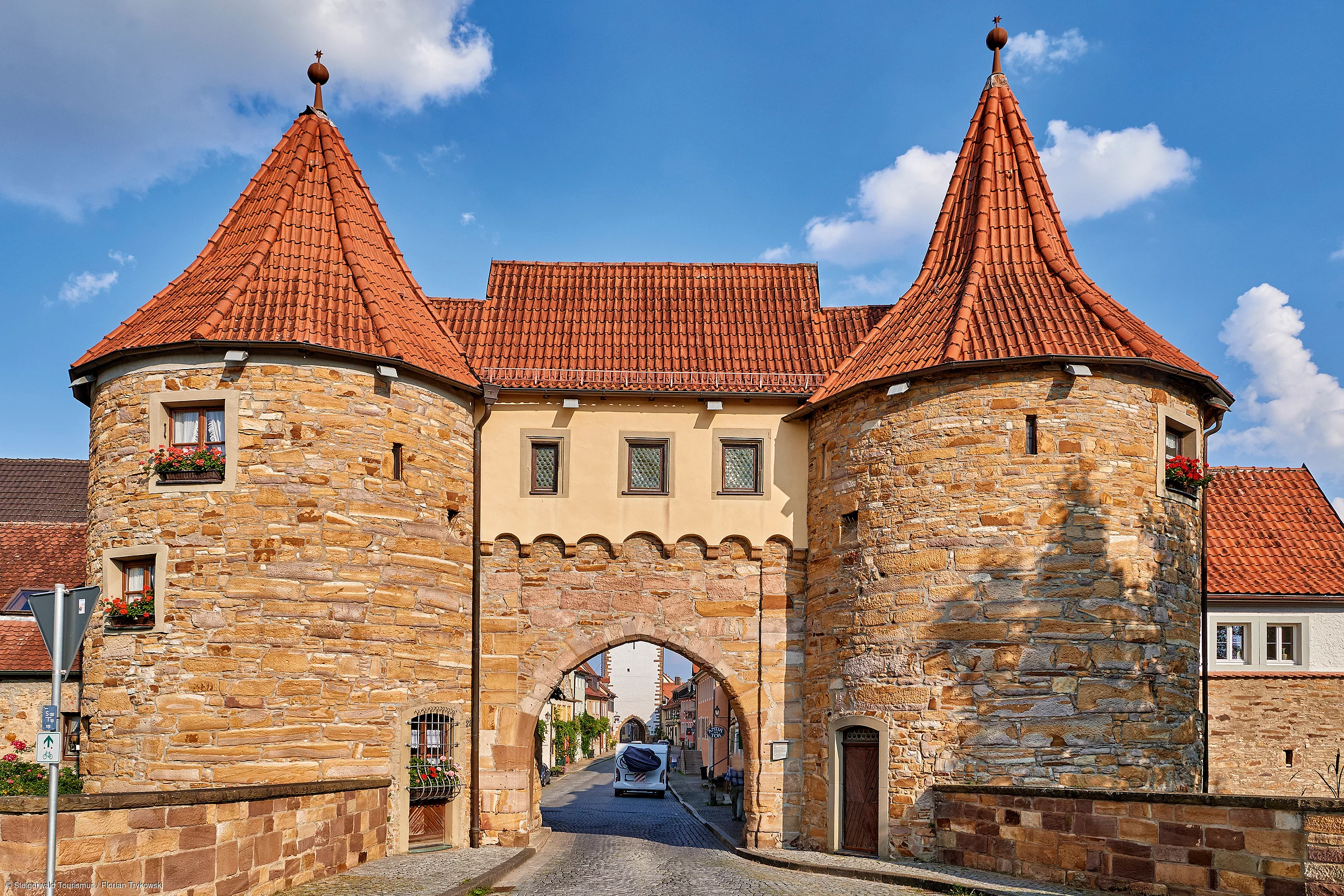 Ein historisches Stadttor mit roten Dächer und zwei runden Türmen an den Ecken gibt den Blick frei hinein in ein mittelalterliches Städtchen