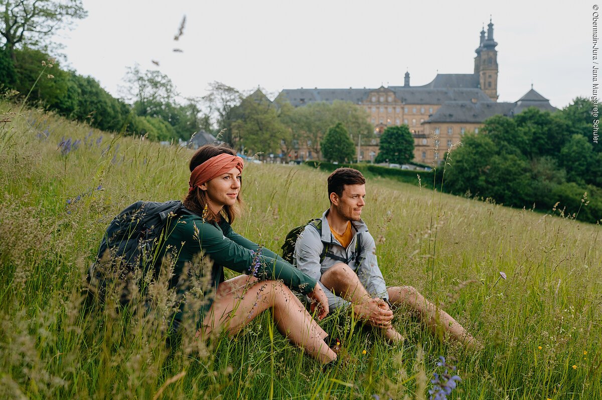 Zwei Wandernde machen Rast auf einer Wiese, im Hintergrund eine Klosteranlage mit Türmen