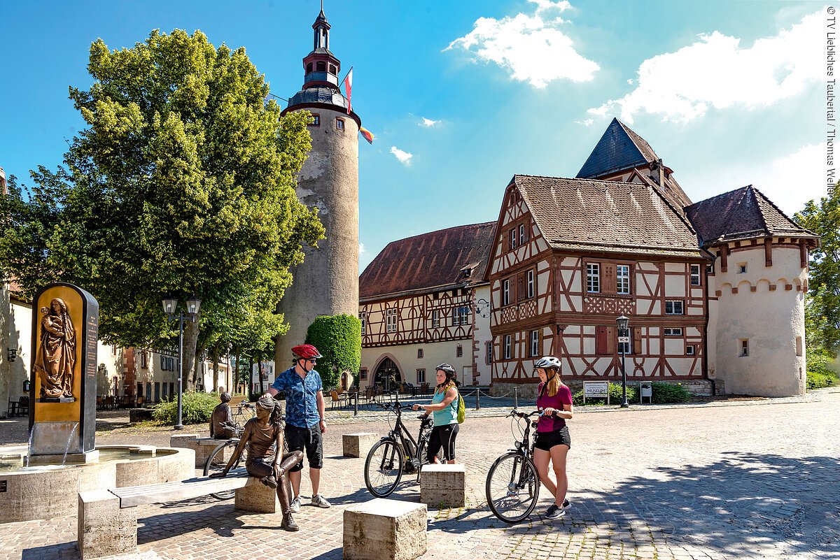 Vor einem historischen Gebäudekomplex mit Fachwerk steht eine Gruppe Radfahrer:innen