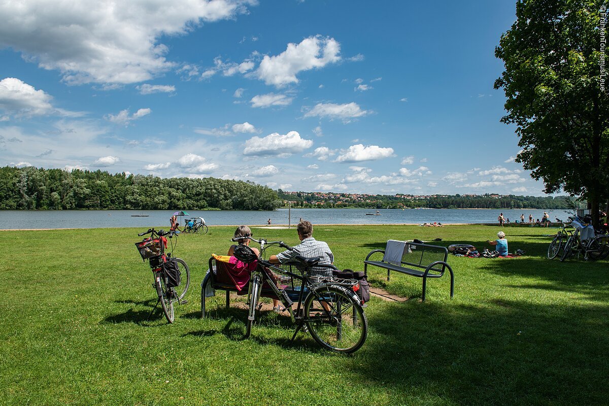 Auf einer Gruppe Bänke inmitten einer Wiese vor einem See machen Radelnde Pause