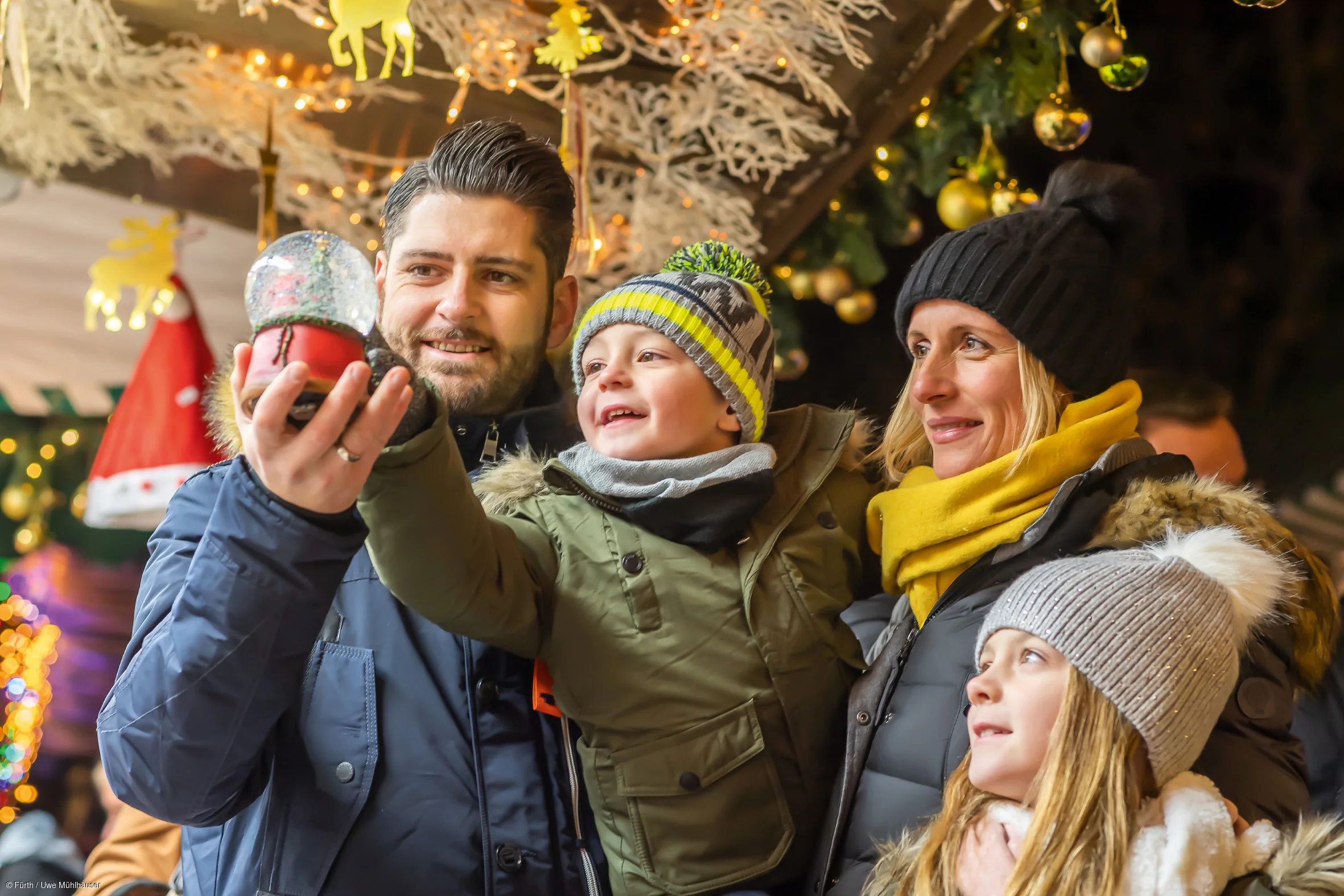 Familie auf einem Weihnachtsmarkt, hält eine Schneekugel. Hintergrund mit Lichtern und Dekorationen.