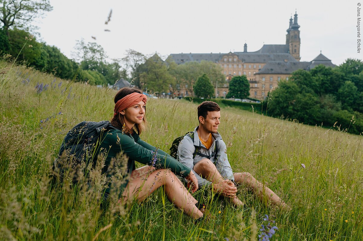 Wandern Kloster Banz (Bad Staffelstein, Obermain-Jura) Zwei Wandernde machen Rast auf einer Wiese, im Hintergrund eine Klosteranlage mit Türmen