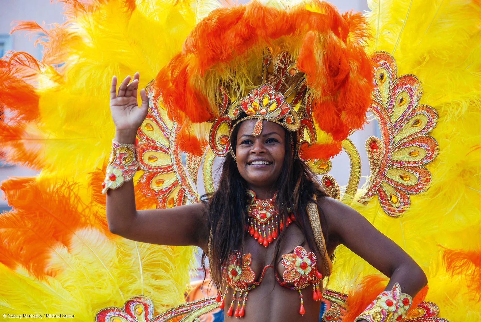 Frau in farbenfrohem Karnevalskostüm mit orangen und gelben Federn und Schmuck tanzt und lächelt.