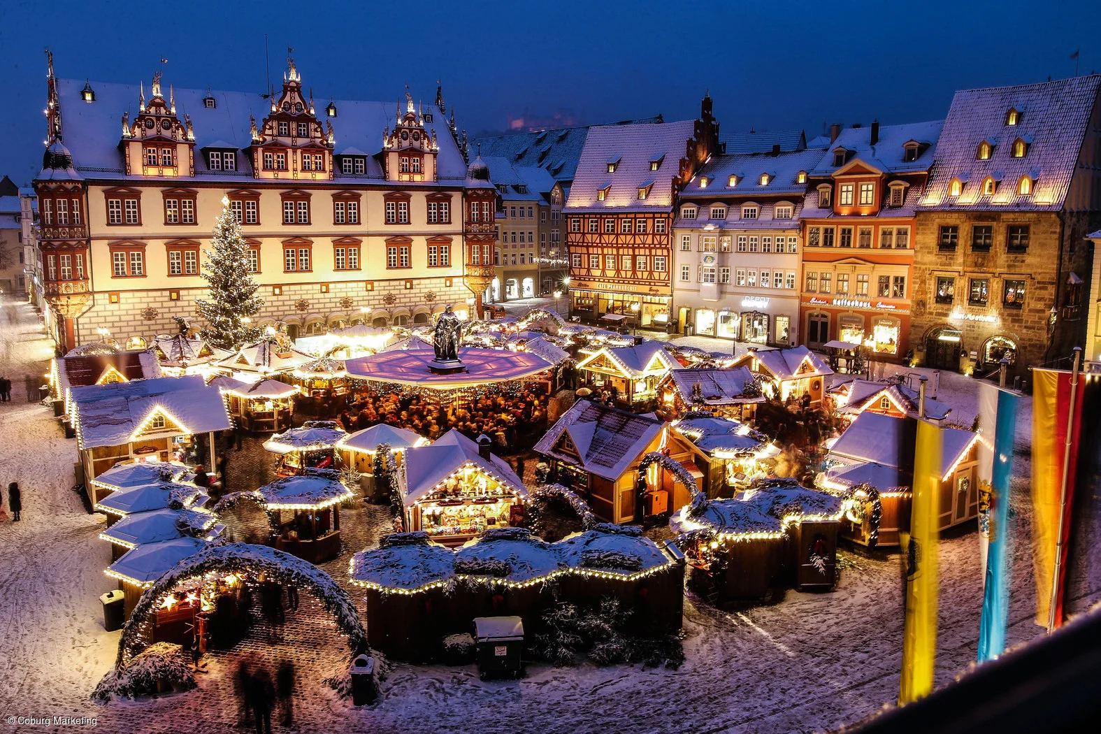 Weihnachtsmarkt mit beleuchteten Ständen und Schnee vor historischen Fachwerkhäusern bei Abenddämmerung.