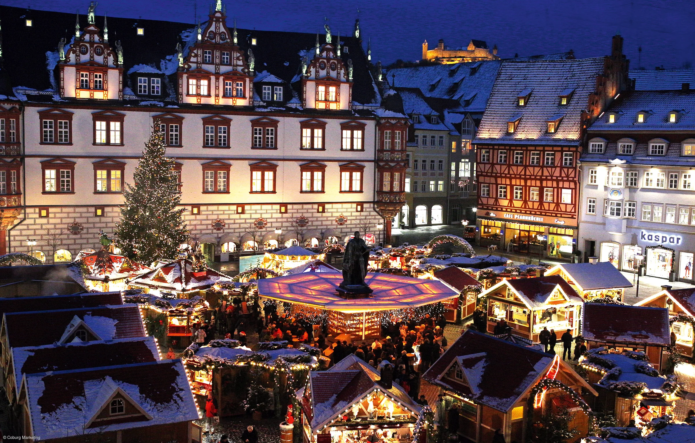 Ein Weihnachtsmarkt mit hell erleuchteten Buden, im nächtlichen Hintergrund eine ebenso illuminierte Burg